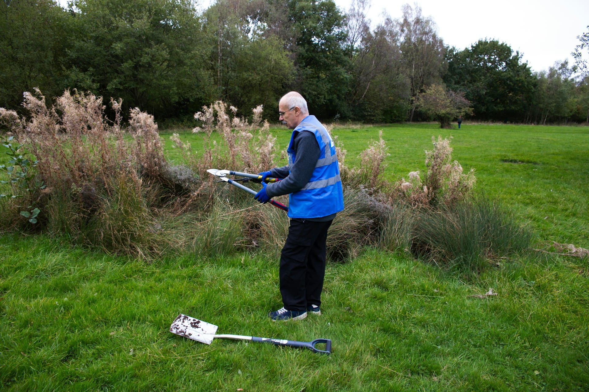 An older man in a hi-vis vest trimming overgrowth during a community conservation task in a park.