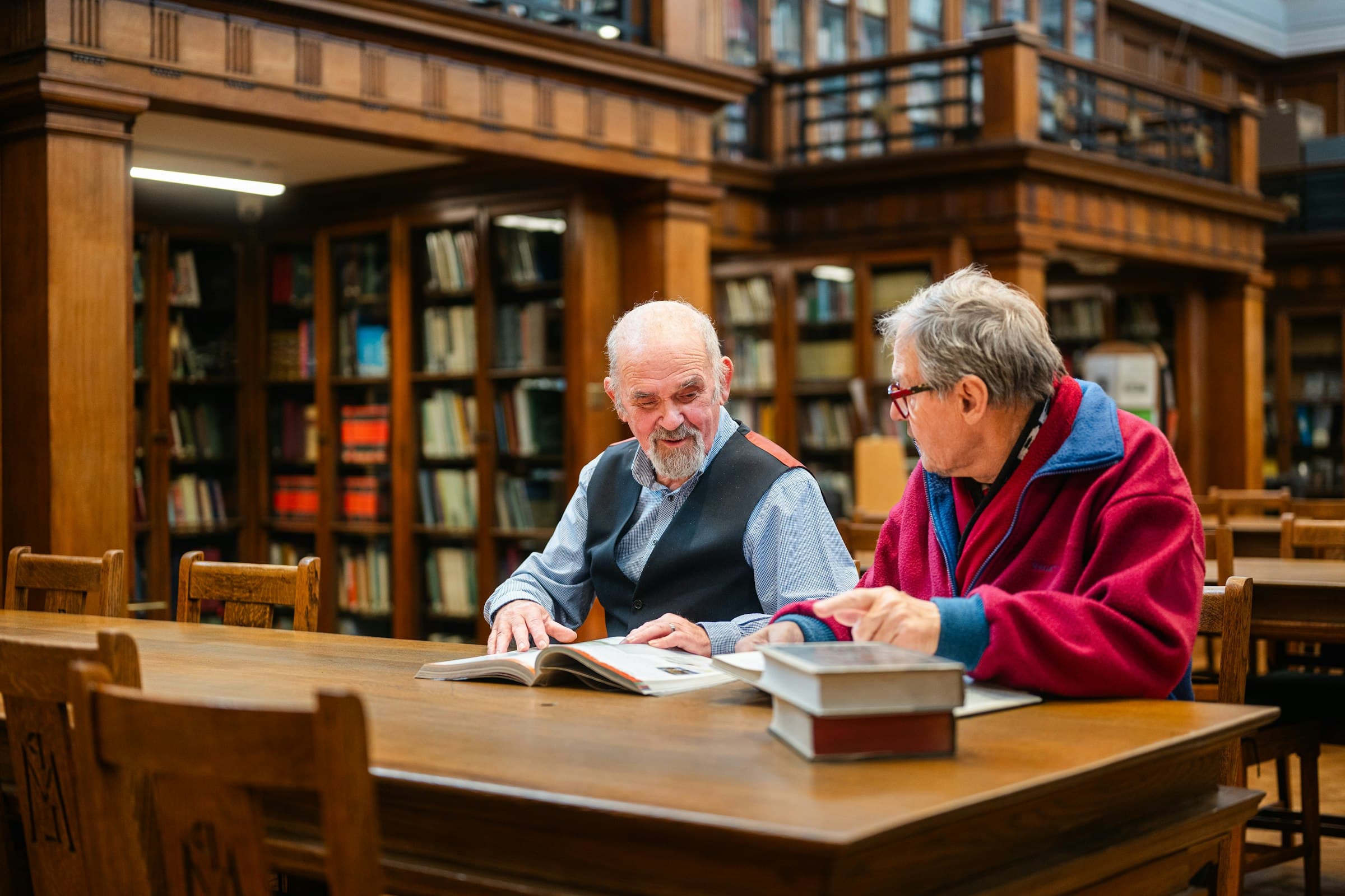 Two older men sat together at a wooden table in a traditional library, talking over an open book.
